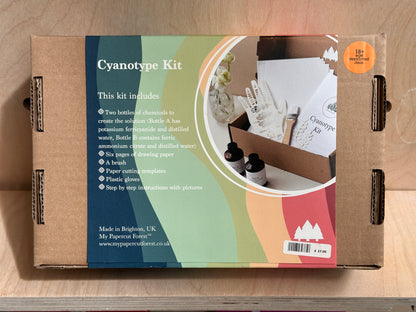 A cardboard box for the Cyanotype Kit by My Paper Forest, photographed from above on a light wood surface. The box features a colorful printed label with text describing the kit contents and a circular photo of the included items.