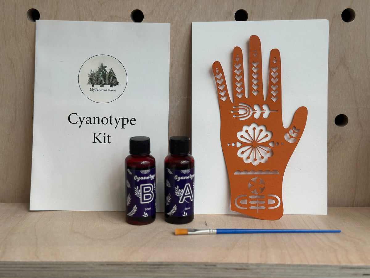 Cyanotype Kit by My Papercut Forests shown with its white booklet, two small bottles of solution, an orange hand-shaped cyanotype plate with floral cutouts, and a paintbrush on a light wood pegboard background.