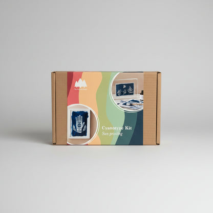 A cardboard box for a cyanotype kit labeled 'My Paper Forest' and 'Cyanotype Kit Sun printing,' shown on a light wood surface. The front of the box has colorful abstract waves and inset photos of blue printed sheets.