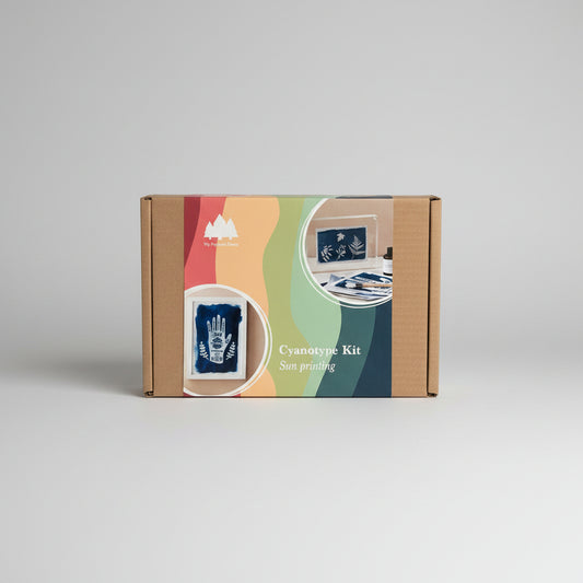 A cardboard box for a cyanotype kit labeled 'My Paper Forest' and 'Cyanotype Kit Sun printing,' shown on a light wood surface. The front of the box has colorful abstract waves and inset photos of blue printed sheets.