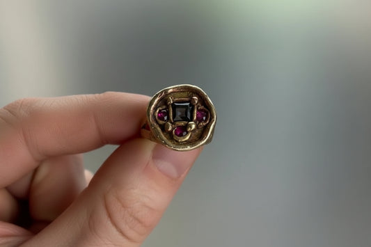 A gold signet ring with a central dark square stone and small purple side stones, held between fingers against a soft gray background.