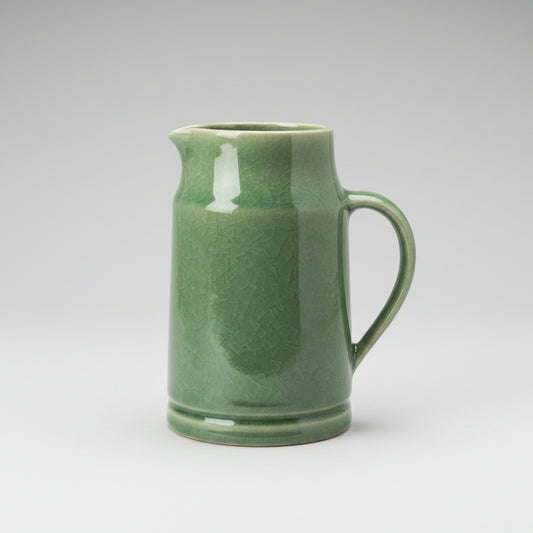 A green ceramic pitcher with a glossy, crackled glaze finish, featuring a spout and a curved handle, set against a plain white background.