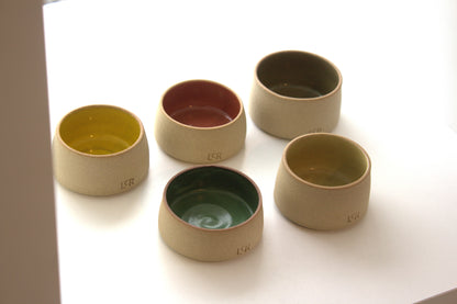 Five small ceramic cups or bowls in a light beige tone, each with a different interior glaze color including yellow, terracotta red, dark green, and olive green, arranged on a white surface.