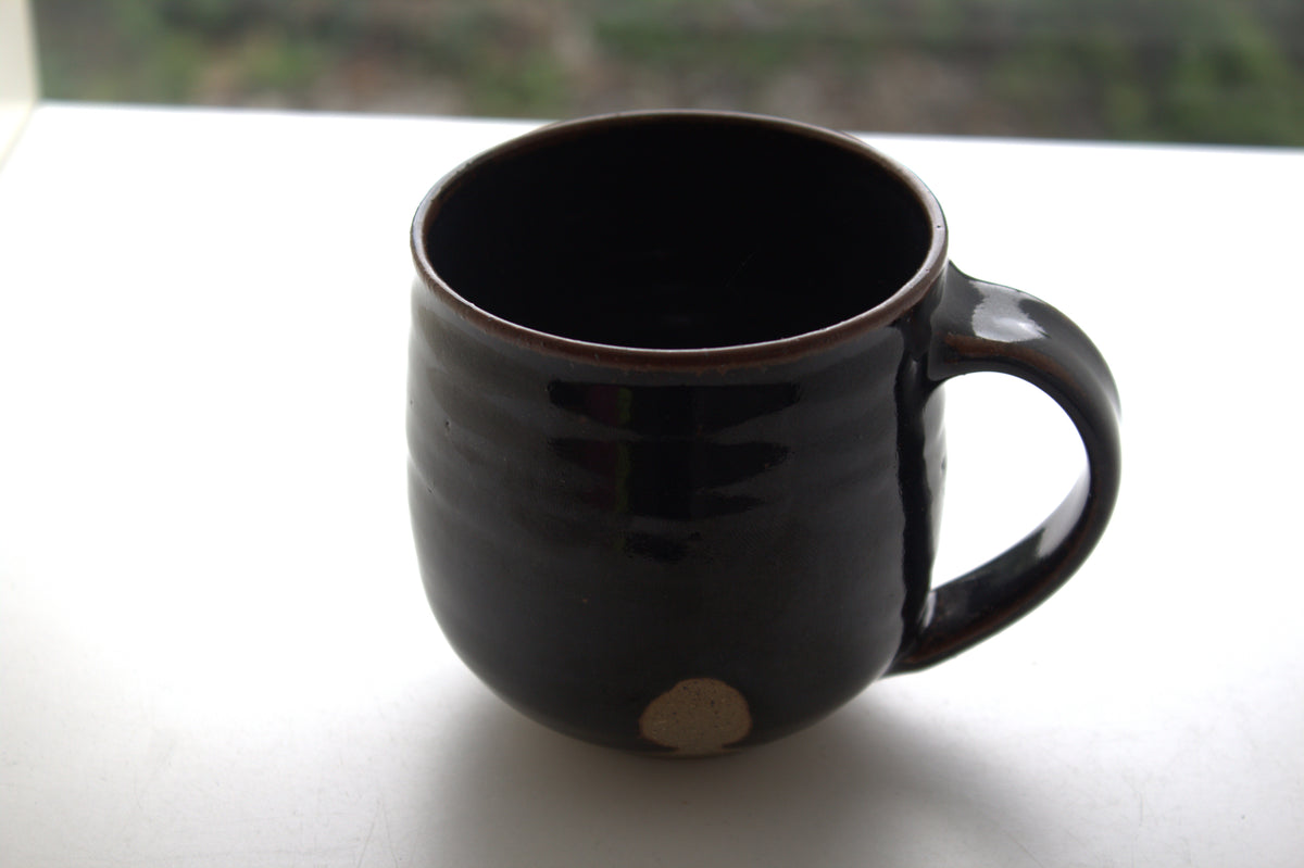 A dark brown glazed ceramic mug with a small beige circular mark near the base, sitting on a white surface with a blurred outdoor background.