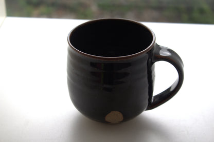 A dark brown glazed ceramic mug with a small beige circular mark near the base, sitting on a white surface with a blurred outdoor background.