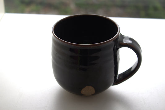 A dark brown glazed ceramic mug with a small beige circular mark near the base, sitting on a white surface with a blurred outdoor background.