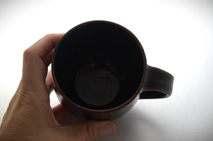 A hand holding a dark brown ceramic mug against a bright white background.