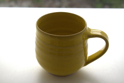 A handmade ceramic mug with a mustard yellow glaze, featuring horizontal ridges and a curved handle, sitting on a white surface.