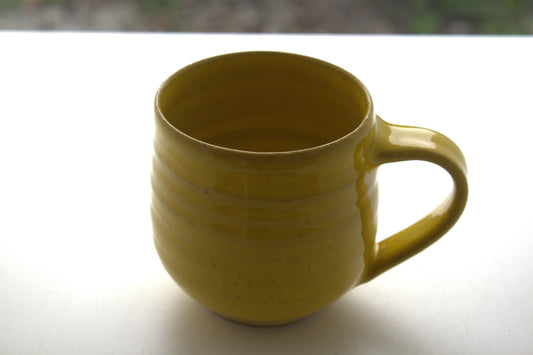 A handmade ceramic mug with a mustard yellow glaze, featuring horizontal ridges and a curved handle, sitting on a white surface.