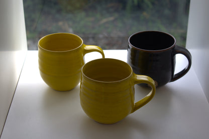 Three ceramic mugs with ribbed textures on a white windowsill, with a yellow mug in front, a second yellow mug behind it, and a dark brown mug to the right. A blurred window view is visible in the background.