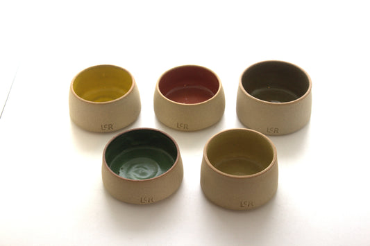 Five small ceramic cups or bowls arranged on a white background, each with a different interior color including mustard yellow, terracotta red, dark green, teal, and light olive. The outer bodies are a warm beige ceramic, and the embossed letters 'LSR' are visible on the front of each cup.