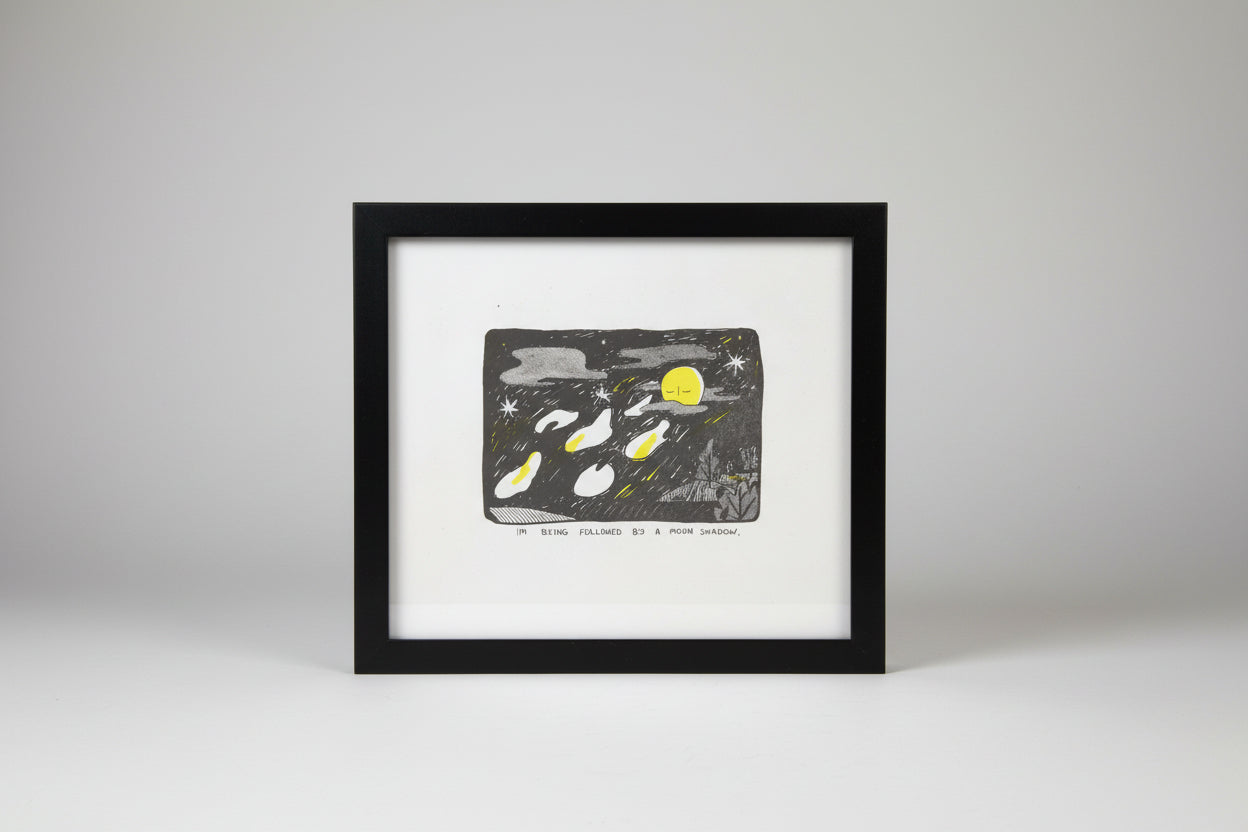 A hand-drawn style art print or card featuring a night sky scene with a yellow moon, stars, and several white and yellow moon phases. Below the illustration, handwritten text reads, 'I'M BEING FOLLOWED BY A MOON SHADOW.
