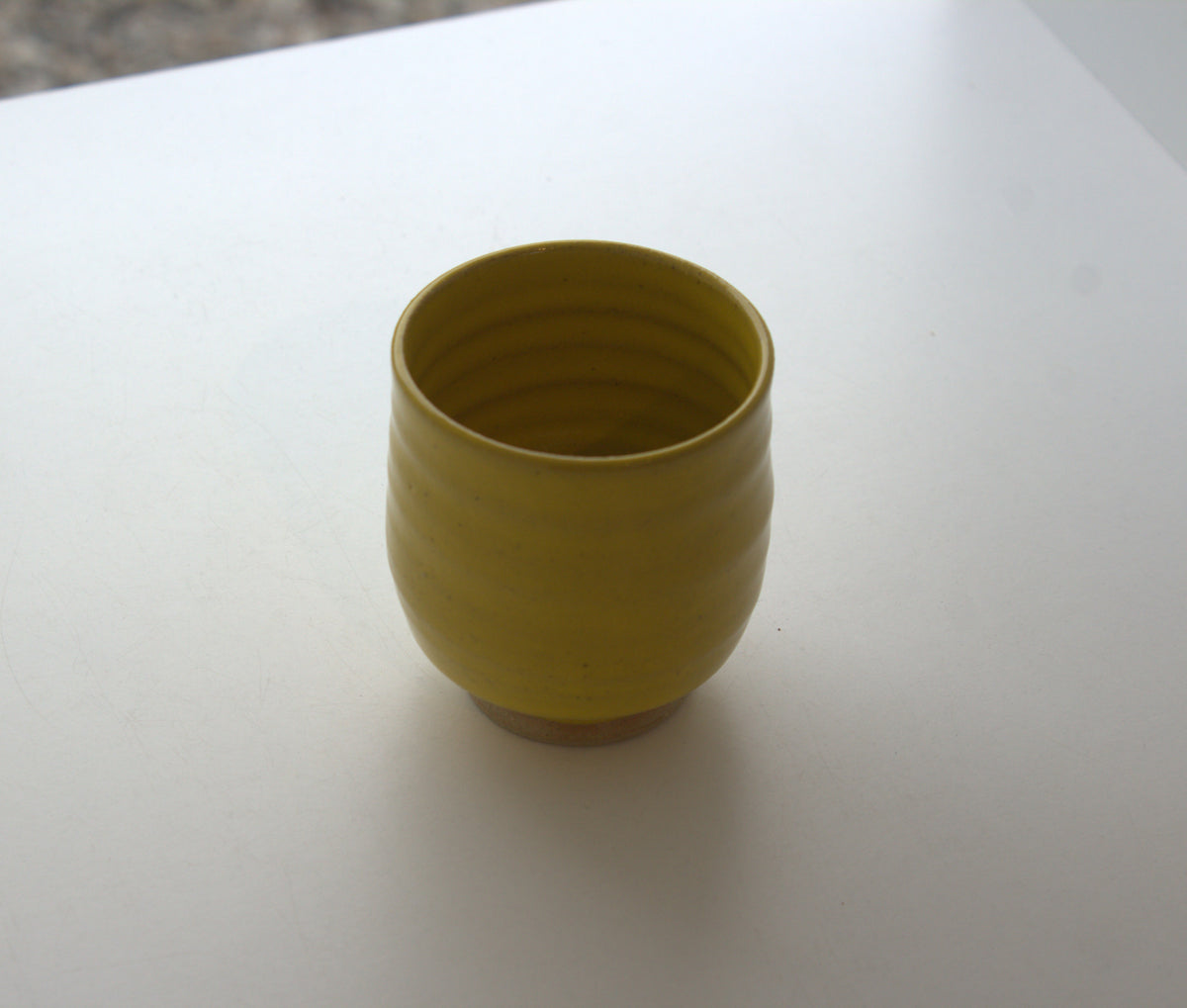 A small yellow ceramic cup with a ribbed exterior and a natural unglazed brown base, sitting on a plain white surface.