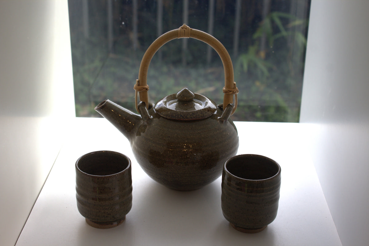 A dark brown ceramic teapot with a light bamboo handle, accompanied by two matching ceramic cups, set on a white surface in front of a window.
