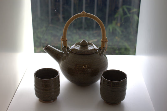 A dark brown ceramic teapot with a light bamboo handle, accompanied by two matching ceramic cups, set on a white surface in front of a window.