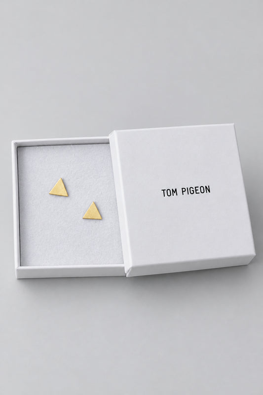 A hand holds an open white TOM PIGEON jewelry box showing two small gold triangle stud earrings against a blurred laptop background.
