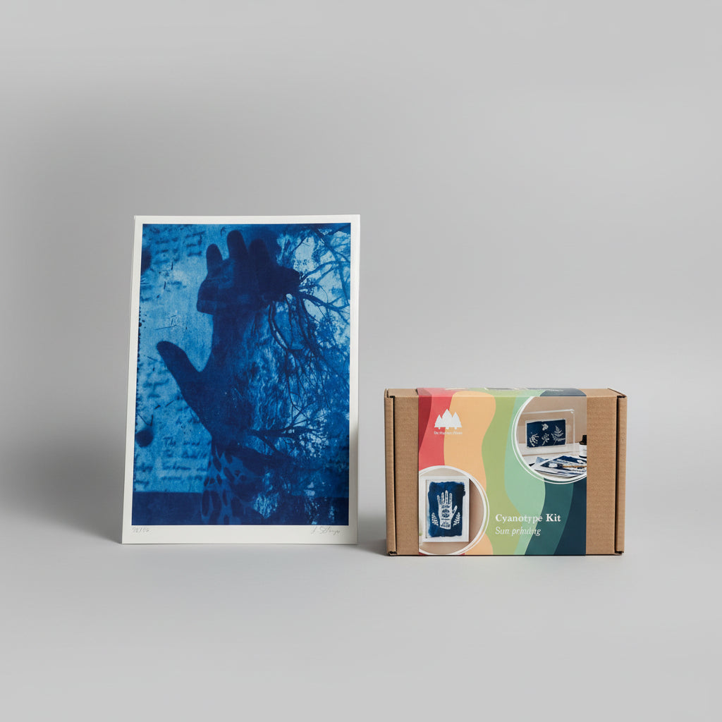 A cyanotype art print with a blue handprint and tree silhouette, displayed next to its cardboard packaging box labeled 'Cyanotype Kit Sun printing' on a wooden pegboard shelf.