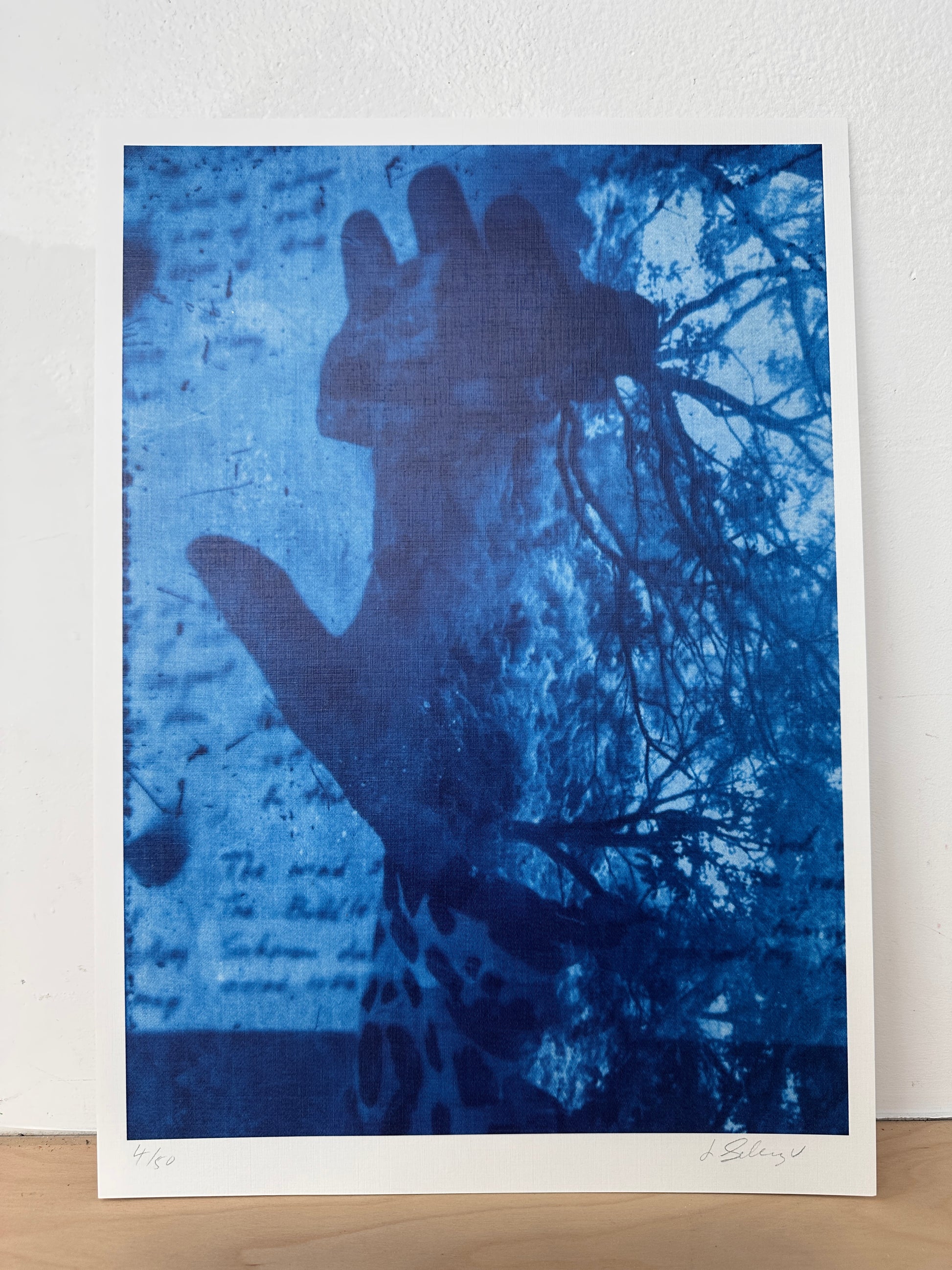 A blue-toned art print or poster leaning against a white wall on a light wood surface. The artwork shows a dark hand silhouette over a textured blue background with script and tree branches.