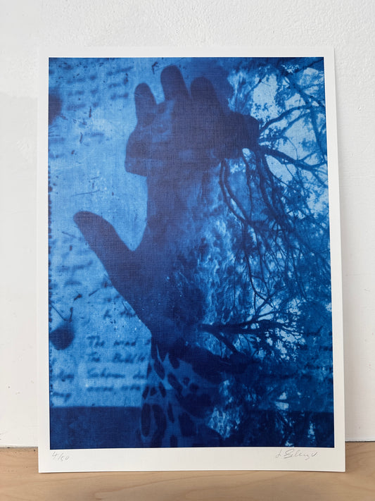 A blue-toned art print or poster leaning against a white wall on a light wood surface. The artwork shows a dark hand silhouette over a textured blue background with script and tree branches.