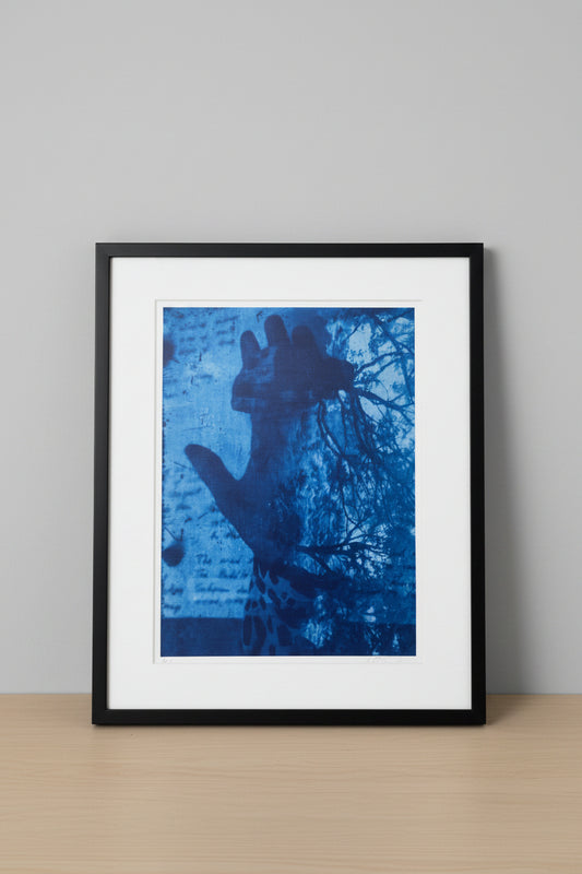 A blue-toned art print or poster leaning against a white wall on a light wood surface. The artwork shows a dark hand silhouette over a blue textured background with tree branches and script-like marks.