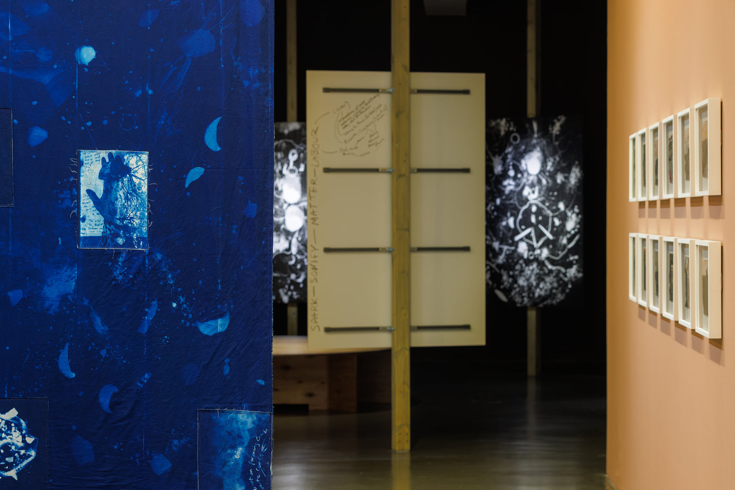 An art gallery interior showing a large textured blue wall installation on the left, a central wooden partition with horizontal bars, glowing black-and-white abstract screens, and a peach-colored wall on the right with multiple framed artworks.