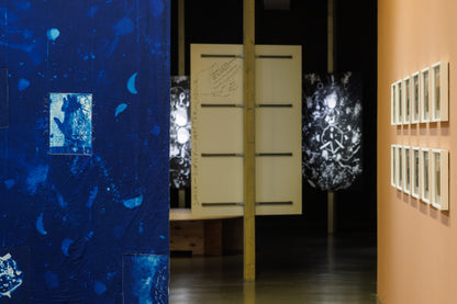 An art gallery interior showing a large textured blue wall installation on the left, a central wooden partition with horizontal bars, glowing black-and-white abstract screens, and a peach-colored wall on the right with multiple framed artworks.