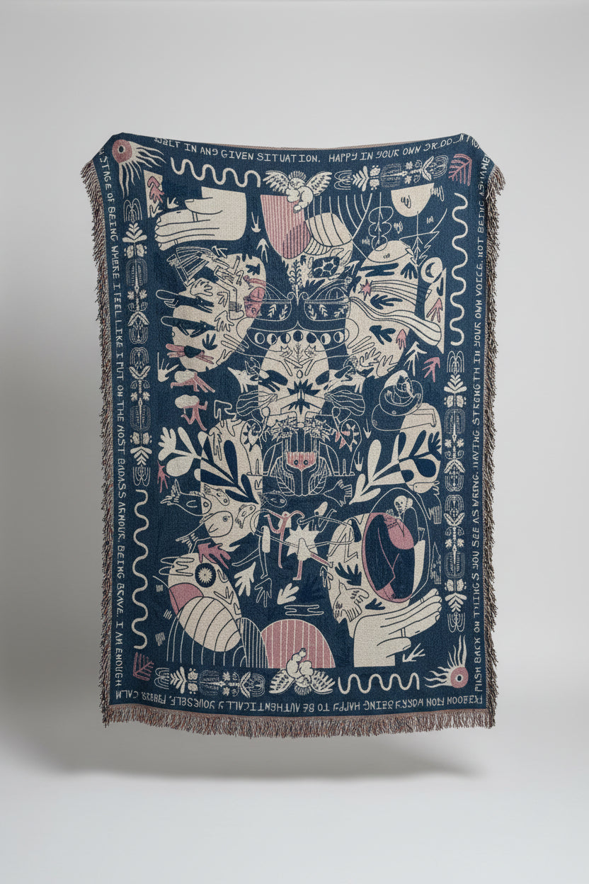 A large woven throw blanket with fringe, featuring a dark blue background and intricate white, pink, and beige illustrated motifs, displayed outdoors against a stone building.