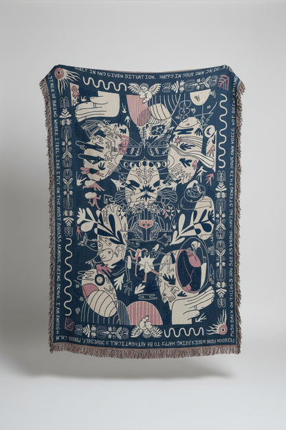 A large woven throw blanket with fringe, featuring a dark blue background and intricate white, pink, and beige illustrated motifs, displayed outdoors against a stone building.