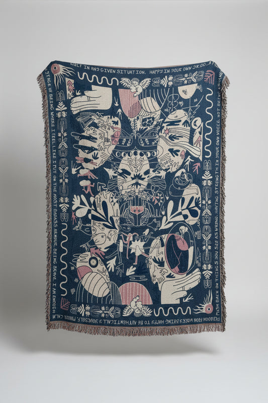 A large woven throw blanket with fringe, featuring a dark blue background and intricate white, pink, and beige illustrated motifs, displayed outdoors against a stone building.